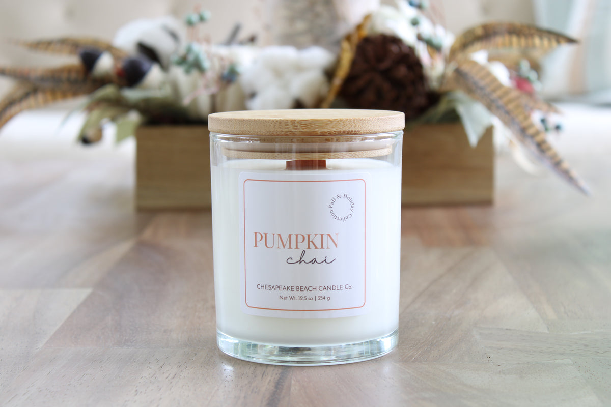 A glass jar candle with a light wood lid and a white label reading 'PUMPKIN chai' sits on a wooden tabletop. The background is softly blurred with autumn decorations, including a wooden box and dried leaves.