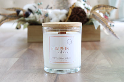 A glass jar candle with a light wood lid and a white label reading 'PUMPKIN chai' sits on a wooden tabletop. The background is softly blurred with autumn decorations, including a wooden box and dried leaves.