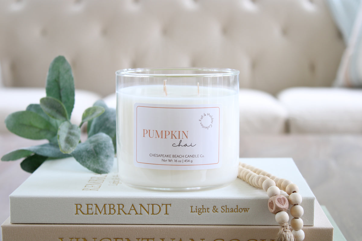 A white soy wax candle in a clear glass jar sits on stacked books, with a green leafy sprig and a wooden bead necklace beside it. The candle label reads 'PUMPKIN CHAI' and shows Chesapeake Beach Candle Co. branding.
