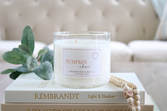A white soy wax candle in a clear glass jar sits on stacked books, with a green leafy sprig and a wooden bead necklace beside it. The candle label reads 'PUMPKIN CHAI' and shows Chesapeake Beach Candle Co. branding.
