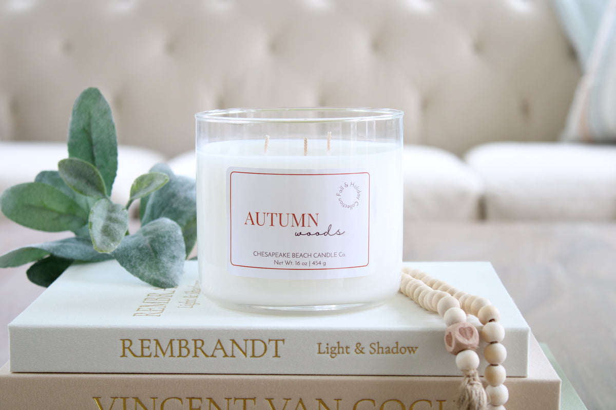 A white candle in a clear glass jar sits on stacked books in a softly styled interior setting. The candle label reads 'AUTUMN woods' and 'Chesapeake Beach Candle Co. 
