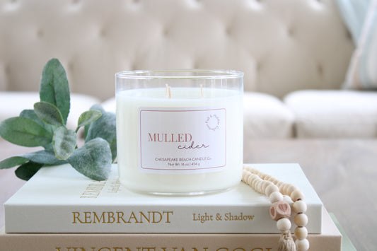 A white soy candle in a clear glass jar sits on stacked books, with a green leafy sprig and a wooden bead garland beside it. The candle label reads 'MULLED CIDER' and the brand appears to be Chesapeake Beach Candle Co.