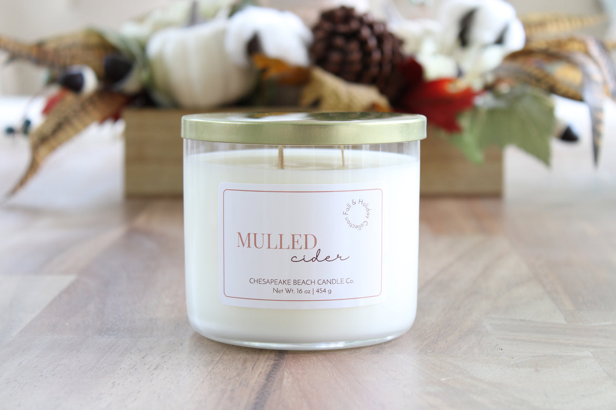 A double-wick candle in a clear glass jar with a gold lid and a white label reading 'MULLED CIDER' by Chesapeake Beach Candle Co., placed on a wooden surface with a blurred autumn-themed background.