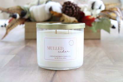 A double-wick candle in a clear glass jar with a gold lid and a white label reading 'MULLED CIDER' by Chesapeake Beach Candle Co., placed on a wooden surface with a blurred autumn-themed background.