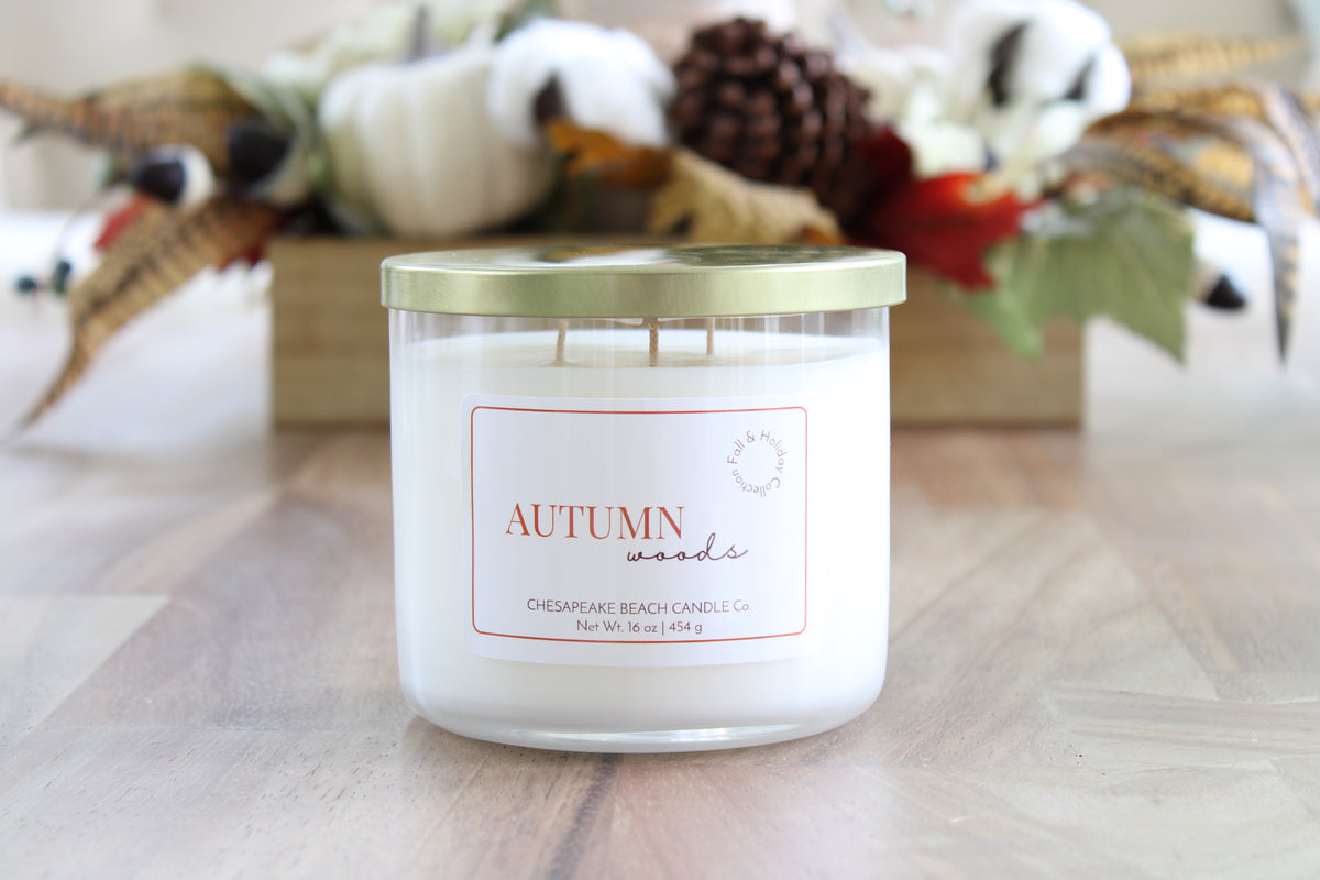 A three-wick soy candle in a clear glass jar with a white wax fill and a metallic gold lid, labeled 'AUTUMN woods' by Chesapeake Beach Candle Co. The candle is placed on a light wood surface with a blurred autumn-themed decorative box in the background.