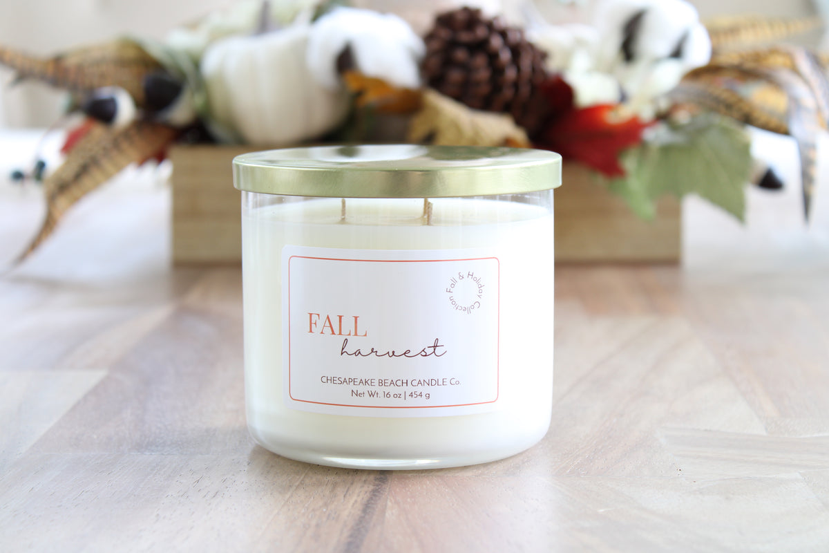 A triple-wick candle in a clear glass jar with a gold lid and a white label reading 'Fall harvest' Chesapeake Beach Candle Co.' It sits on a light wood surface with a blurred autumn-themed background featuring pumpkins, cotton bolls, and leaves.