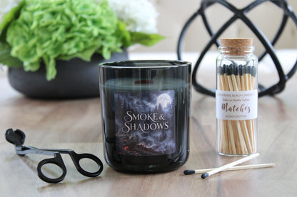 A dark glass candle jar labeled 'Smoke & Shadows' sits on a wooden surface, styled with a black wick trimmer, a small glass jar of matchsticks, and a blurred green plant in the background.